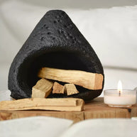 Black natural stone incense burner filled with palo santo wood pieces, displayed on a wooden tray with a lit tealight candle against a soft neutral fabric background
