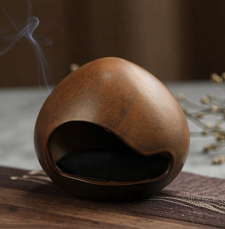 Side view of a handmade brown-glazed ceramic incense burner shaped like a yawning face, with incense smoke curling above it.