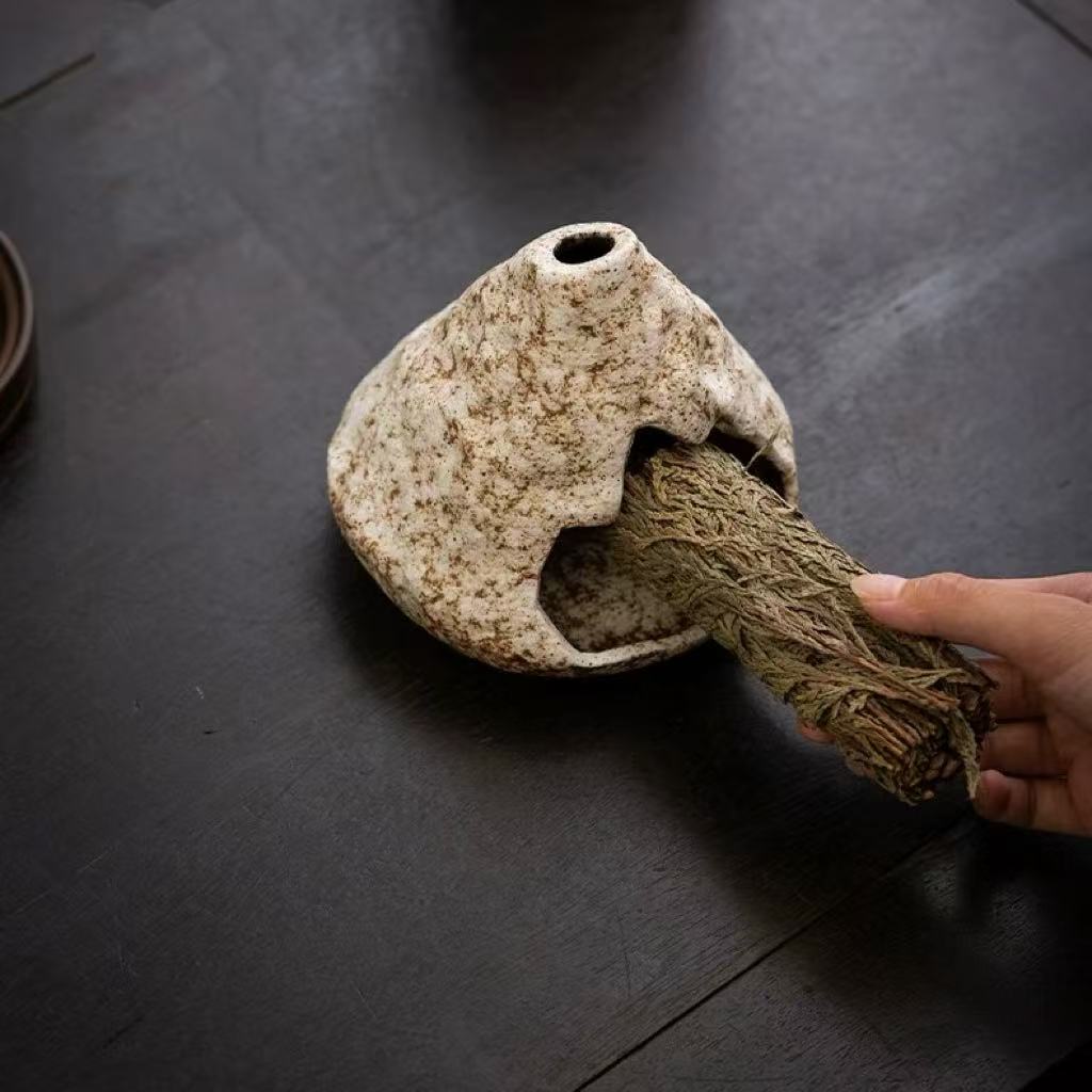 Handmade ceramic incense burner with a sage smudge stick being placed inside, displayed on a dark textured surface for a rustic aesthetic