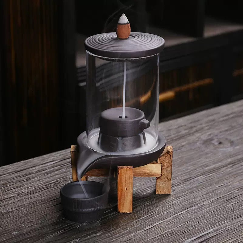 Modern glass backflow incense burner with metal base and wooden stand, placed on a wooden tabletop.