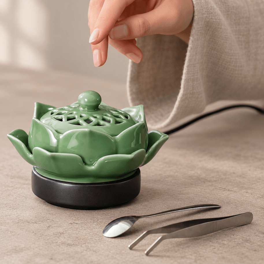 A hand above the decorative lid of a green ceramic lotus electric incense burner, with a spoon and tweezers beside it.