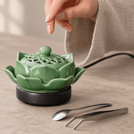 A hand above the decorative lid of a green ceramic lotus electric incense burner, with a spoon and tweezers beside it.