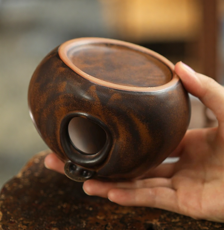 Hand holding a handmade brown-glazed ceramic incense burner, showing the unglazed flat base and the side opening.