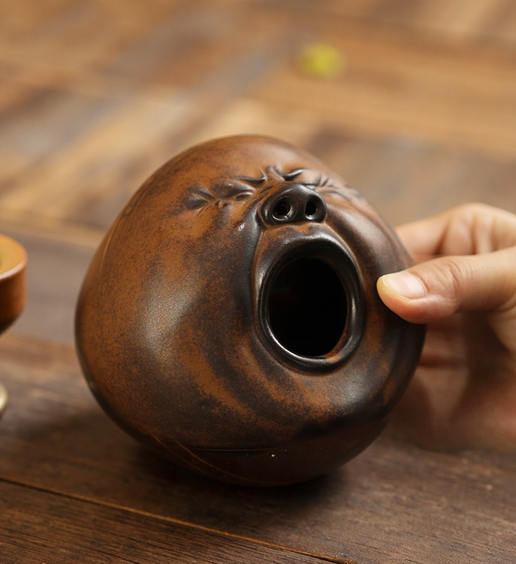 A hand holding a handmade brown-glazed ceramic incense burner shaped like a yawning face, showing the wide mouth opening and nostril vents