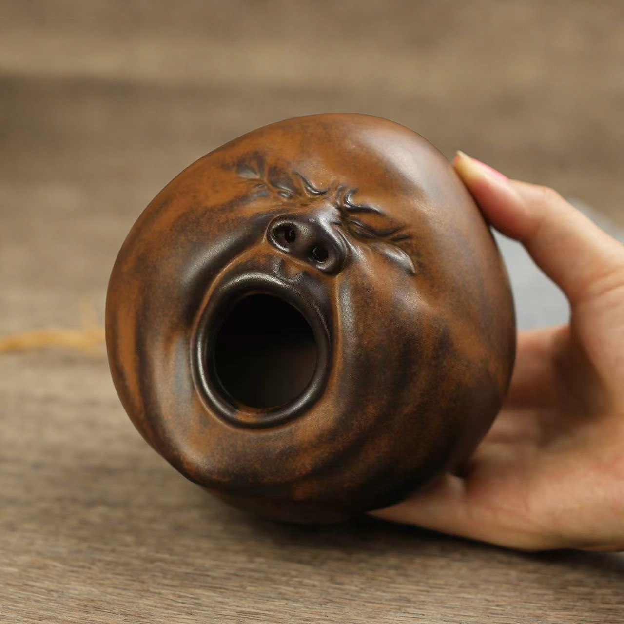 Close-up of a hand holding a handmade brown-glazed ceramic incense burner shaped like a yawning face.
