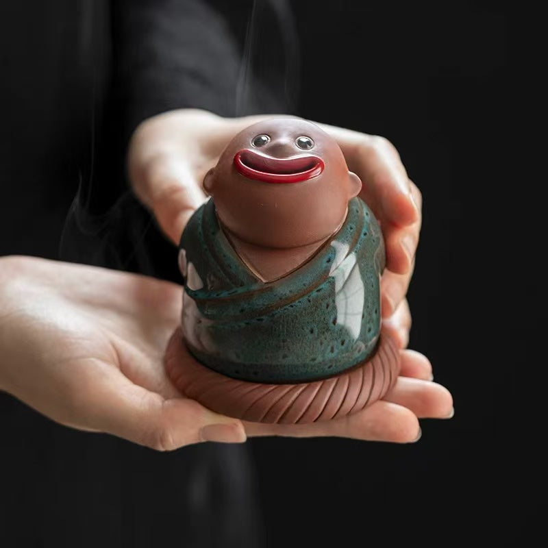 Two hands gently holding a green ceramic monk incense burner with a smiling face