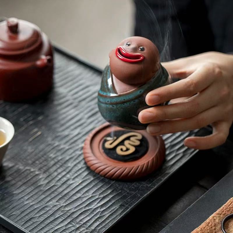 Hand holding a green-robed ceramic monk incense burner with smoke flowing from its mouth
