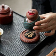 Hand holding a green-robed ceramic monk incense burner with smoke flowing from its mouth
