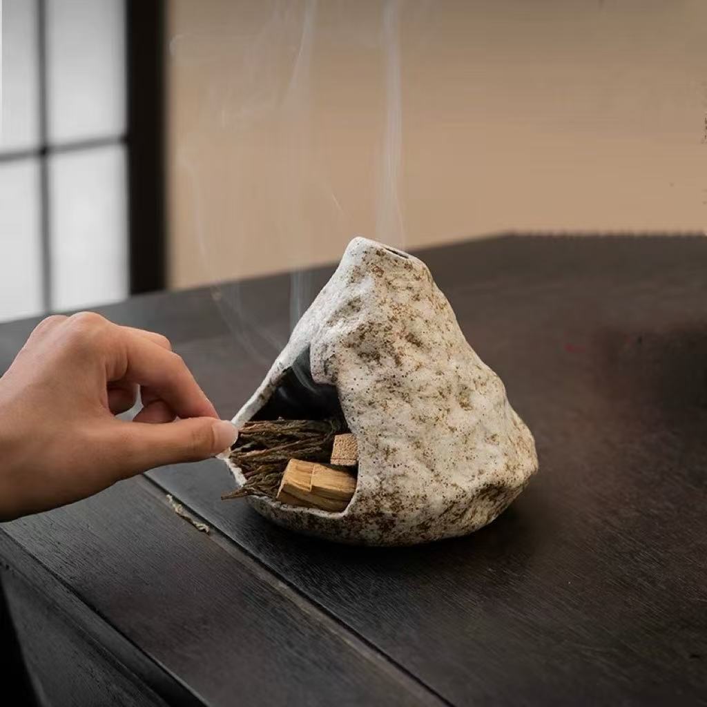 Hand lighting a bundle of natural incense inside a handmade ceramic incense burner with visible smoke, placed on a dark wooden table in a traditional room setting