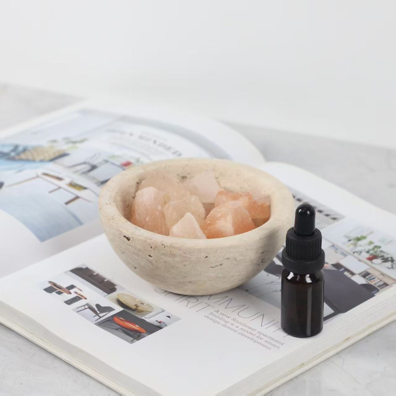 Natural travertine bowl filled with pink Himalayan crystals next to essential oil bottle, styled on an open magazine