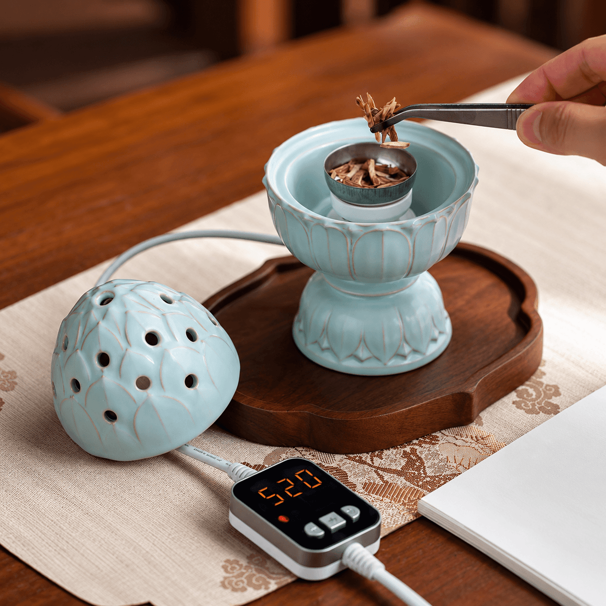 Hand using tweezers to add incense chips to a pale blue lotus electric burner, with the lid removed and controller in front.