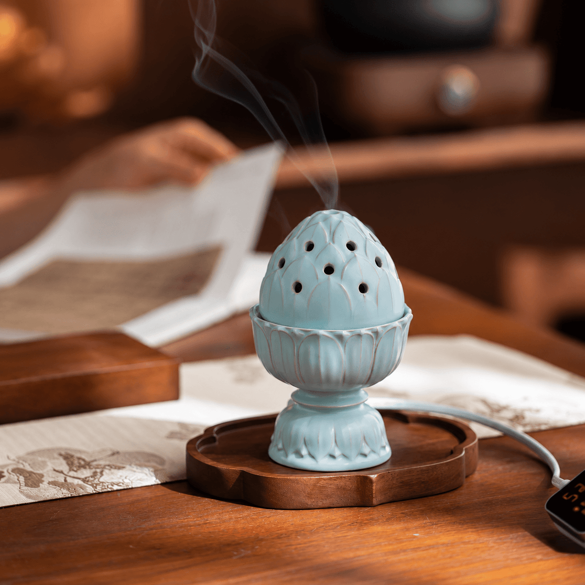Pale blue lotus electric incense burner on a wooden tray, with smoke rising from the lid and someone reading in the background.