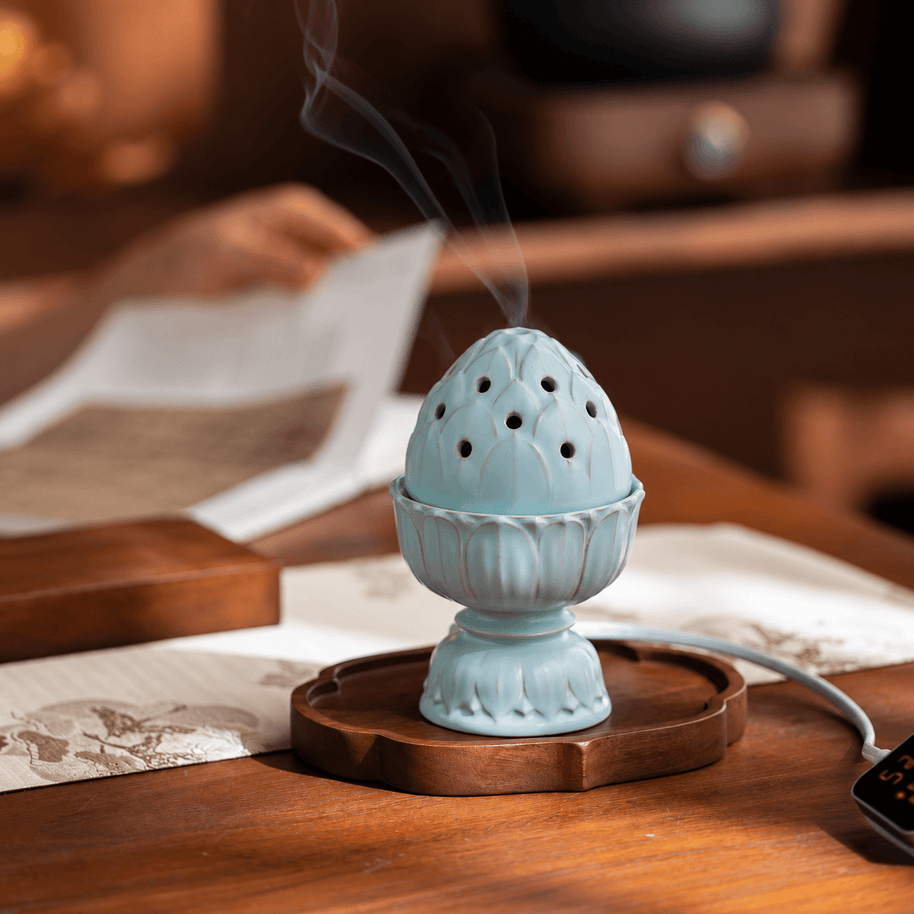 Pale blue lotus electric incense burner on a wooden tray, with smoke rising from the lid and someone reading in the background.