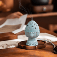 Pale blue lotus electric incense burner on a wooden tray, with smoke rising from the lid and someone reading in the background.