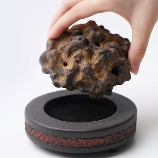 Hand holding the top piece of a detachable rock-form purple clay incense burner with red-patterned base on white background