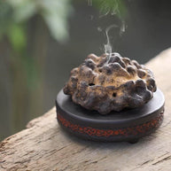 Rock-form purple clay incense burner emitting smoke on wooden surface with green nature background
