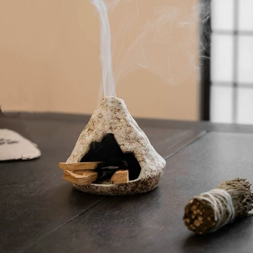 Handmade ceramic incense burner emitting vertical stream of fragrant smoke, with sandalwood chips burning inside and a sage smudge stick placed beside on a dark table