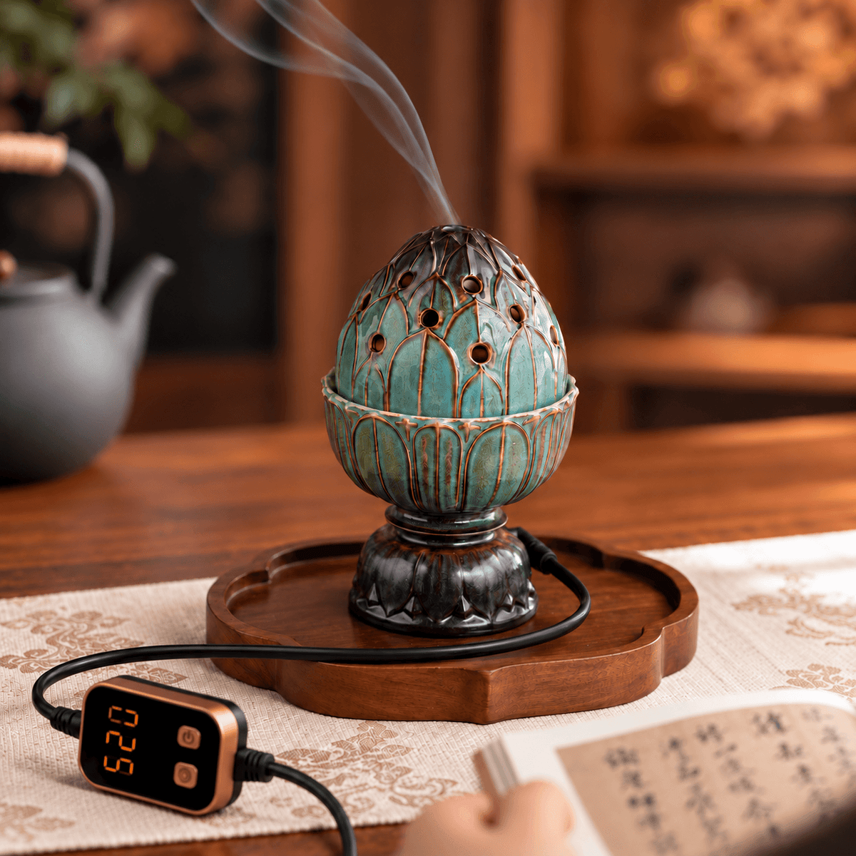 Teal lotus electric incense burner releasing smoke on a wooden tray beside an open book, with a digital controller in front.