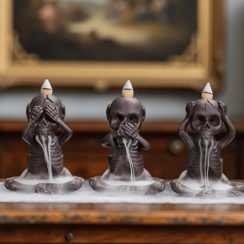 Set of three skeleton-shaped backflow incense burners in "hear no evil, see no evil, speak no evil" poses with smoke flowing from their mouths.