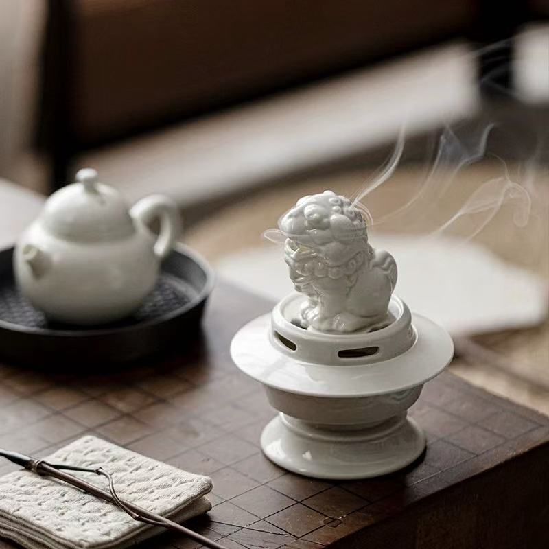 White ceramic lion incense burner releasing smoke on a wooden tea table with teapot and incense tools in background
