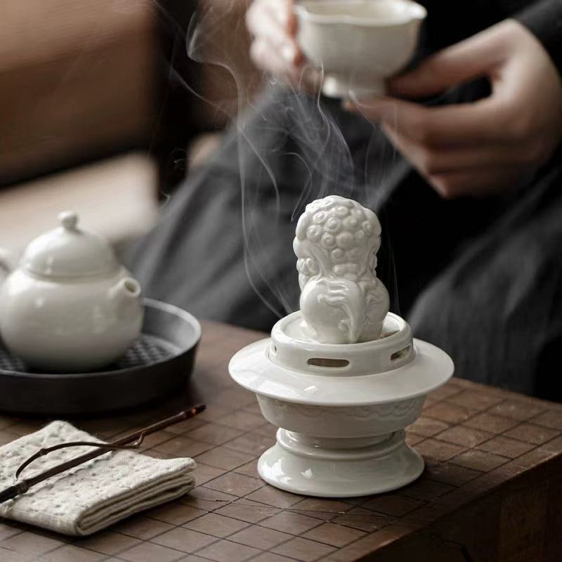 White ceramic Suanni lion incense burner emitting smoke on a tea table with a person holding a teacup in the background
