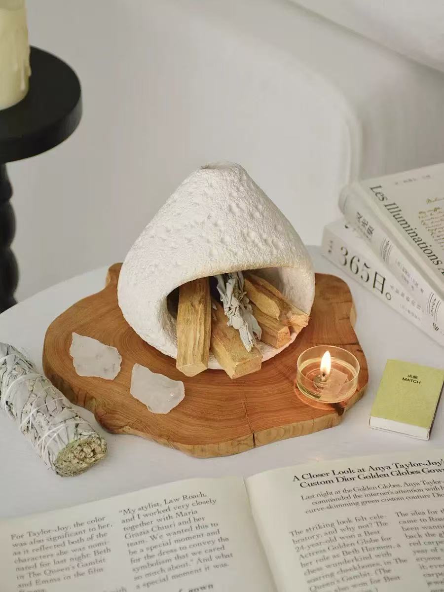 White natural stone incense burner on a carved wooden tray, surrounded by palo santo sticks, white sage bundle, raw crystals, open books, and a lit tealight candle in a calming spiritual setup