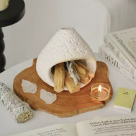 White natural stone incense burner on a carved wooden tray, surrounded by palo santo sticks, white sage bundle, raw crystals, open books, and a lit tealight candle in a calming spiritual setup