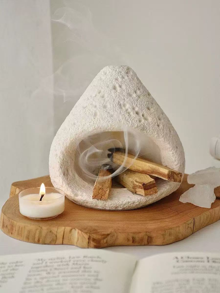 Natural white stone incense burner with burning incense emitting spiral smoke, placed on a carved wooden tray with a tealight candle and open book in a peaceful setting