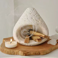 Natural white stone incense burner with burning incense emitting spiral smoke, placed on a carved wooden tray with a tealight candle and open book in a peaceful setting