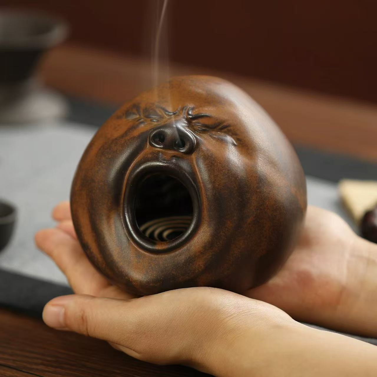 Aromatic ceramic yawning face incense burner held in both hands, with visible spiral incense burning inside